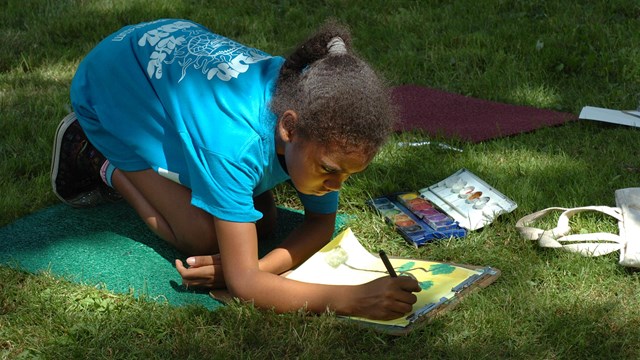 A group of children sitting on mats, painting in a field.