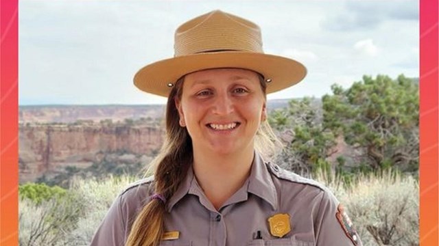 A ranger in front of a grassy landscape with a large red rock canyon.