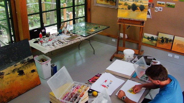 A modern artist's studio with paintings, and tables of paint. An artist works on a pastel drawing.