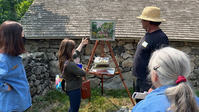 Artists surround a field easel with painting discuss techniques 