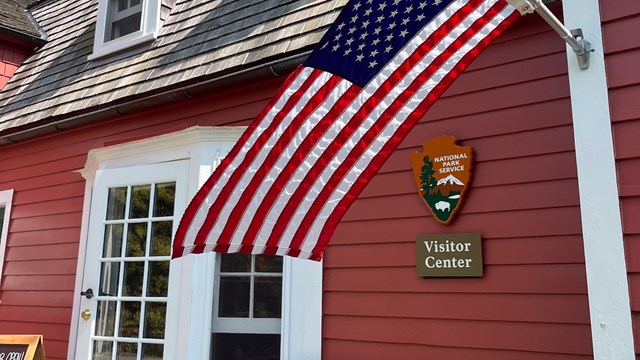 A red historic building with a white door. An American flag flies next to an arrowhead. 