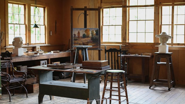 A room with various tables, statue busts, and tools. Bright windows line the walls.