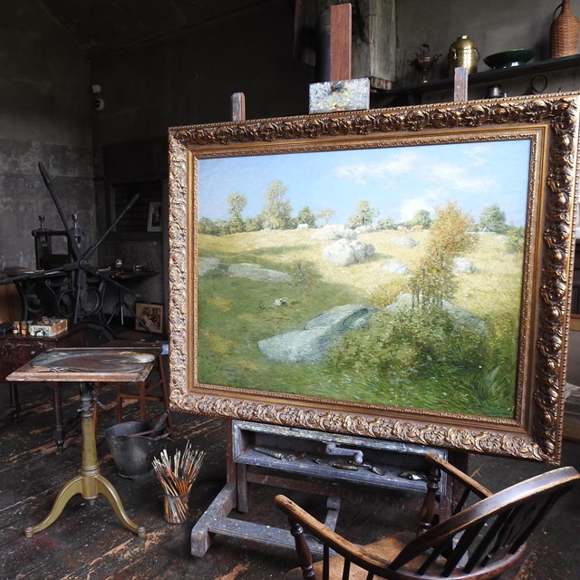 A large painting of a green landscape in a small room filled with easels, chairs, and art supplies.