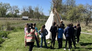 A ranger stands in front of a partially assembled tipi with school kids in front of them