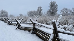 A wooden beam fence and the surrounding area tall grass prairie is covered in snow.