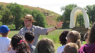 A ranger points to his left at a covered wagon; several school kids are facing the ranger