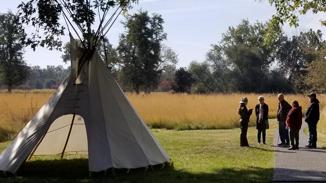 A tipi is set up on the left side of the image and a group of adults on its right in discussion