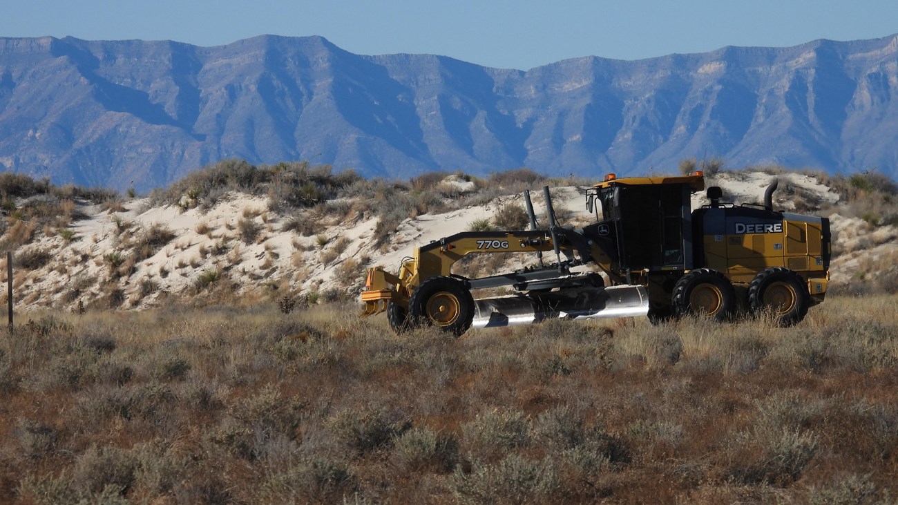 A large yellow "sand plow" shaping the road through an interdunal, vegetated area.