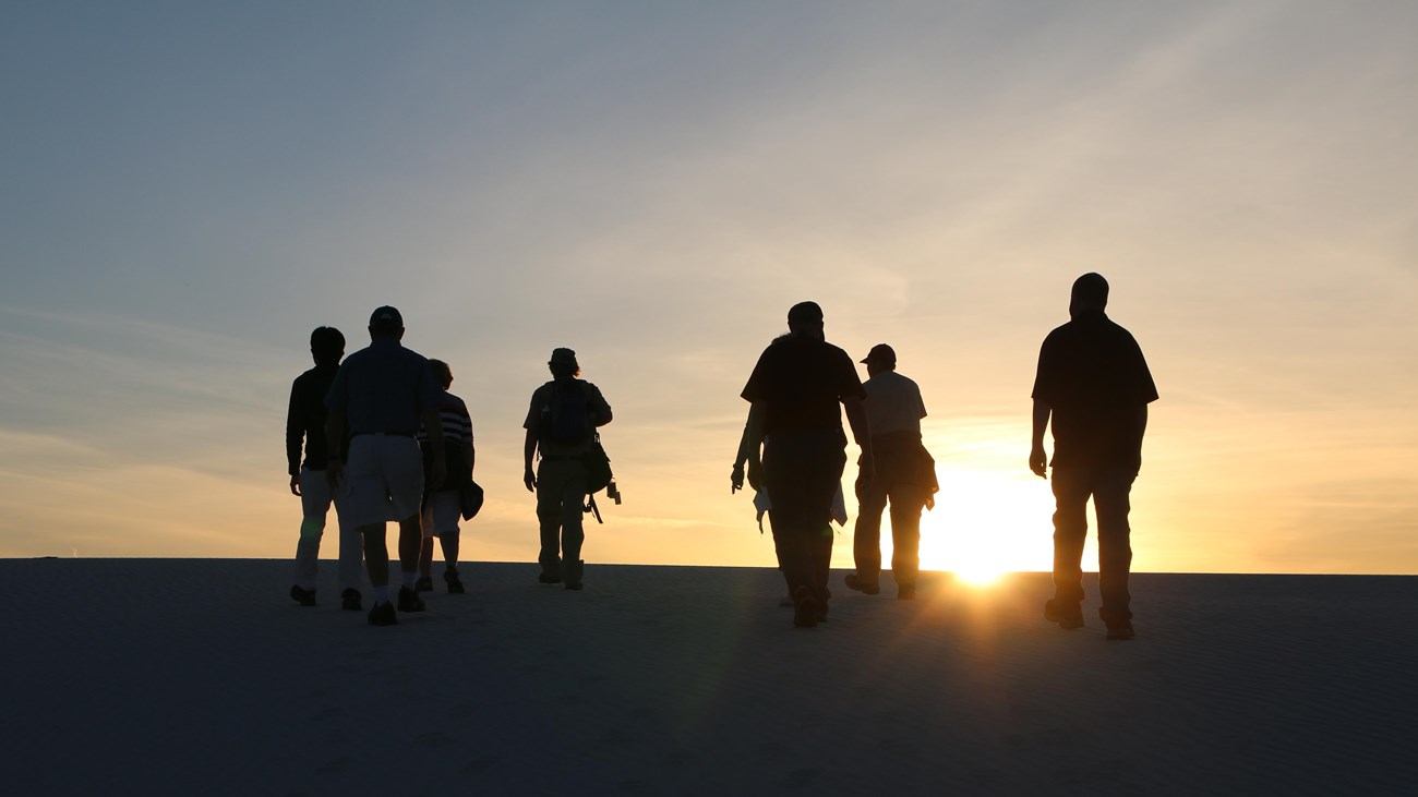 nine people silhouetted by the setting sun as they hike up a white sand dune.