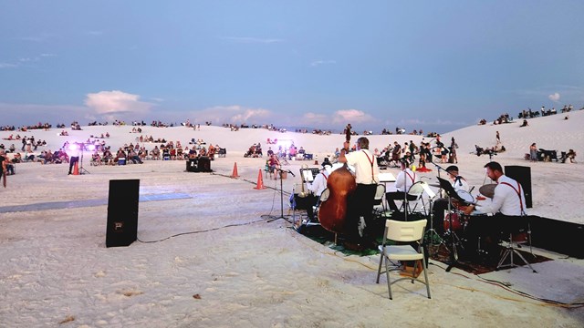 a large group of people in the distance sit on chairs and blankets surrounding six musicians.