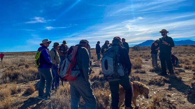 a ranger and a group of people stand on shrubby ground underneath a blue sky. 