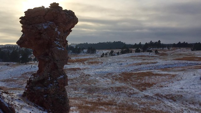 a pillar of brown rock protruding from a hillside overlooking snowy prairie at dusk
