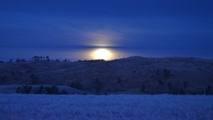 the moon shines through clouds over a frosty, deep blue evening prairie landscape