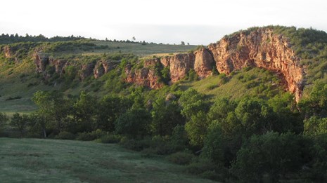 a sharp red cliff side stretching along a green riparian valley at sunset
