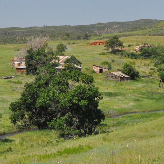 several old wood buildings in a green grassy valley next to a small stream with a few trees
