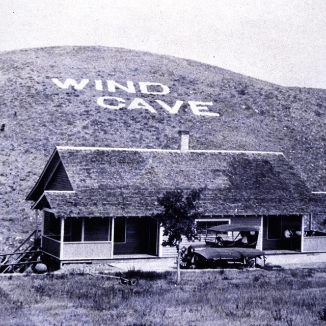 old photo of a building with the American flag and Wind Cave spelled out on the hill behind it
