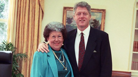 President Clinton stands next to a White woman in an office. His hand rests on her shoulder.