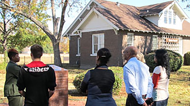 Several people stand in in front of a two story home and listen to a park ranger.