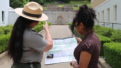 A park ranger points to a fountain in the distance while a woman stands next to her holding a map. 