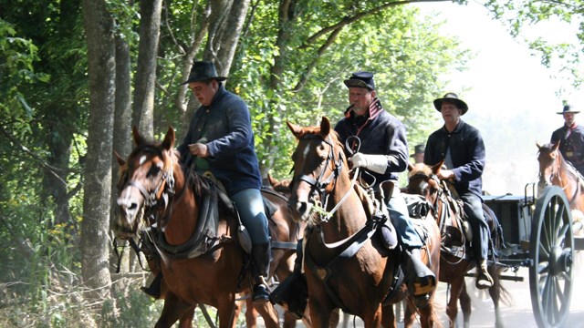 Men wearing Civil War uniforms ride horses as they pull artillery. 