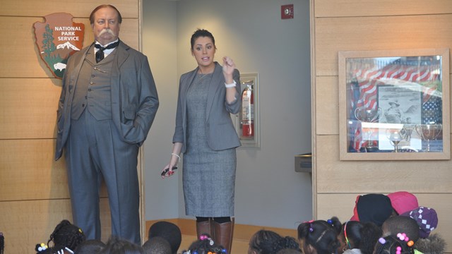 A wax model of a large man in a suit and tie next to a lady in a gray suit speaking to kids