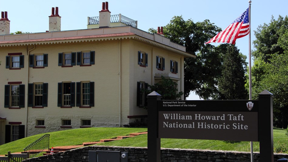 A tall three-story house with green shutters with an American flag and park sign in front