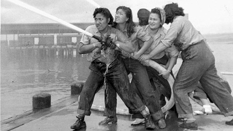 Five women work together to hold fire hose at end of pier