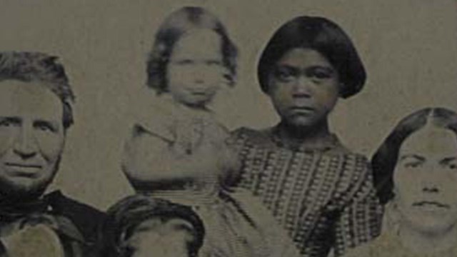 Young indigenous girl in frock holds white baby and looks at camera, surrounded by white family
