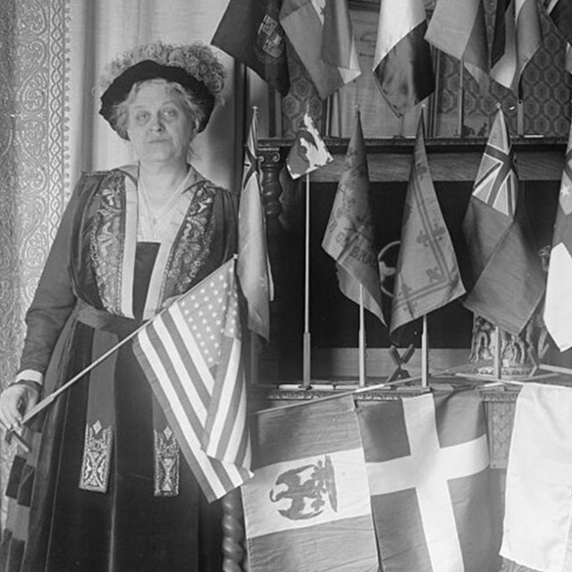 Black and white photo of Carrie Catt holding a US flag in front of many other flags
