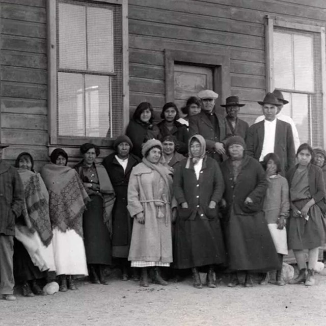 Group of Native Americans stand in front of simple wooden building.