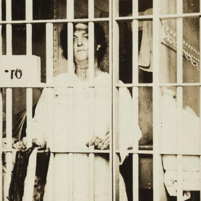 Photograph of Helena Hill Weed, facing forward, standing behind bars in a prison cell. LOC photo