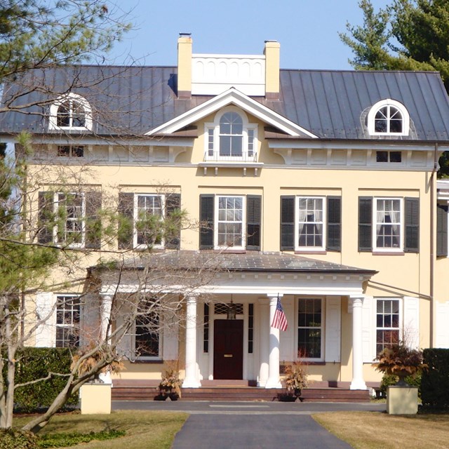 Exterior of a grand home, painted yellow with a grey roof. CC BY SA Zeete Wikimedia
