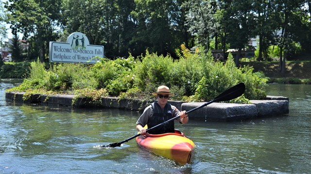 A ranger paddling a kayak past a sign for Seneca Falls