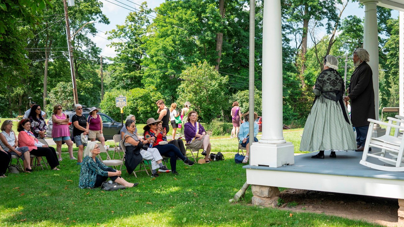 A man and woman in 19th century clothes stand on a porch and address a crowd.