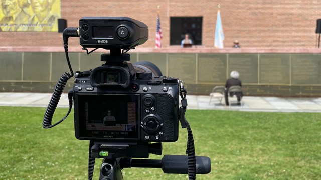 A camera set up outdoors, filming a woman standing in front of a building.