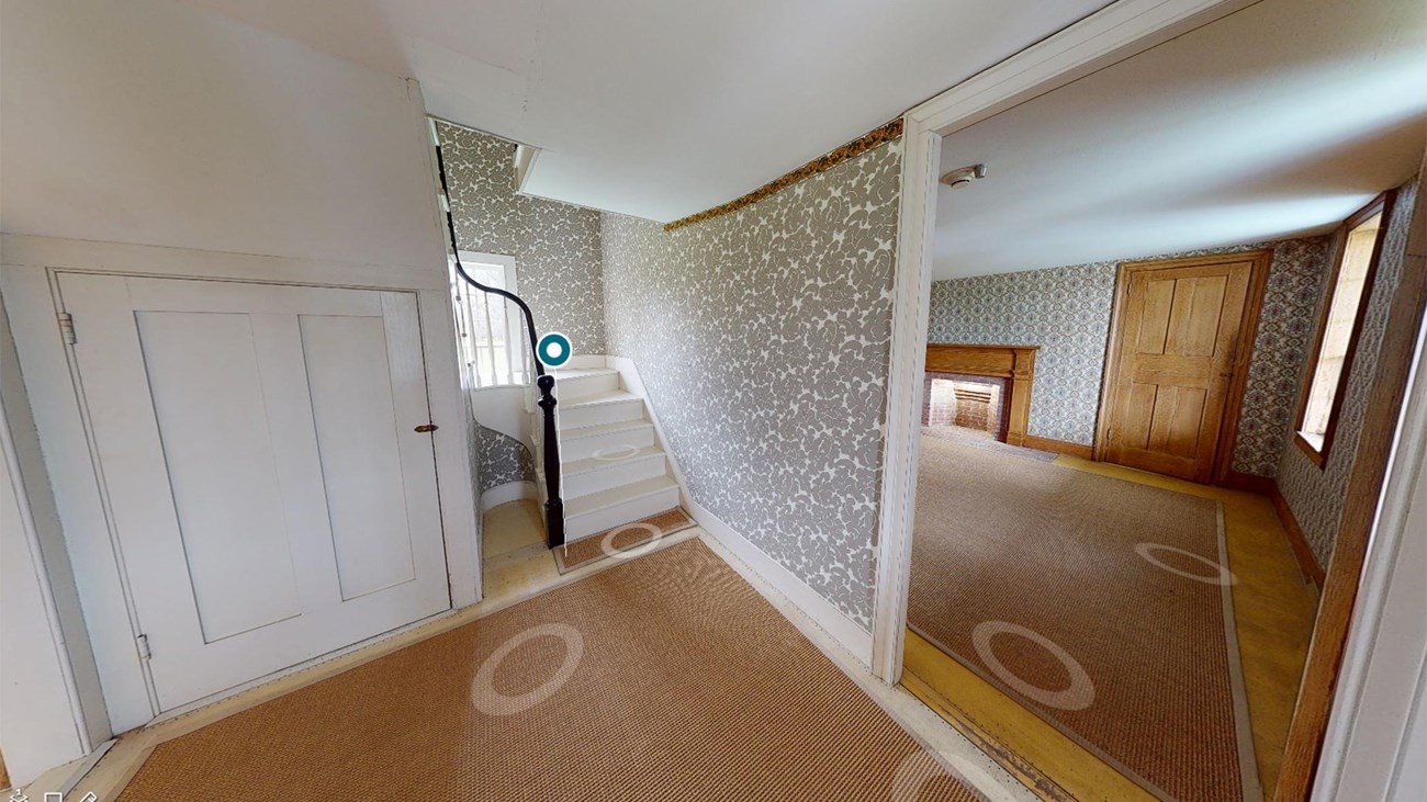 A wide-angle image showing the entryway of a house, including a view of a parlor and a staircase.