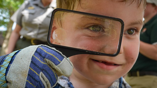 Face of a child with short blond hair looking closely through a magnifying glass at the camera.