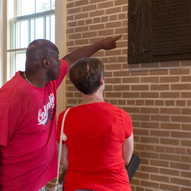 A man and a woman in red shirts look at a plaque on a brick wall, the man points to it.