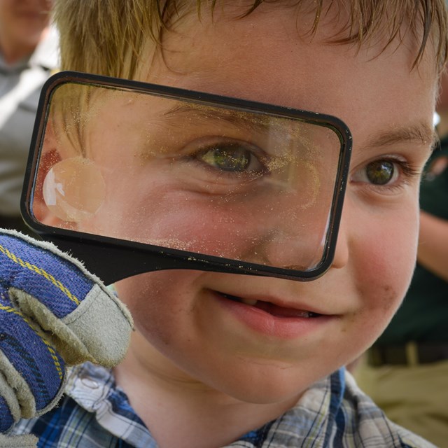 Face of a child with short blond hair looking closely through a magnifying glass at the camera.