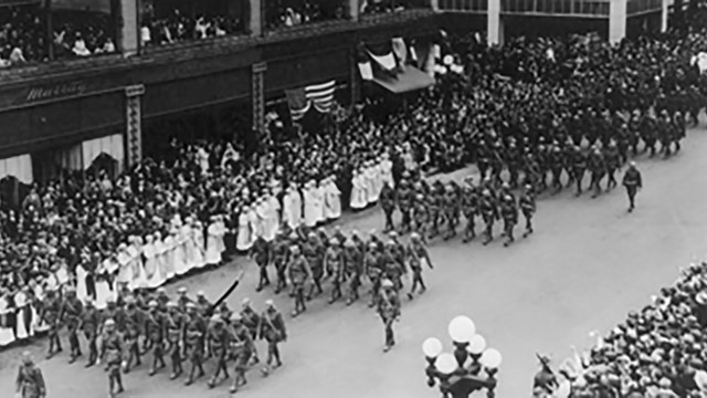 Soldiers march in a parade on a city street lined with spectators.