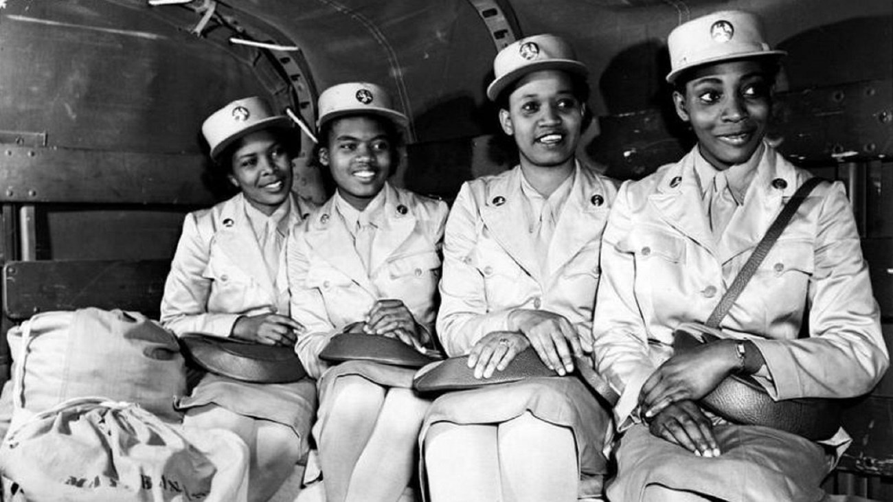 four women wearing military dress uniforms sitting in the back of a troop transport truck.