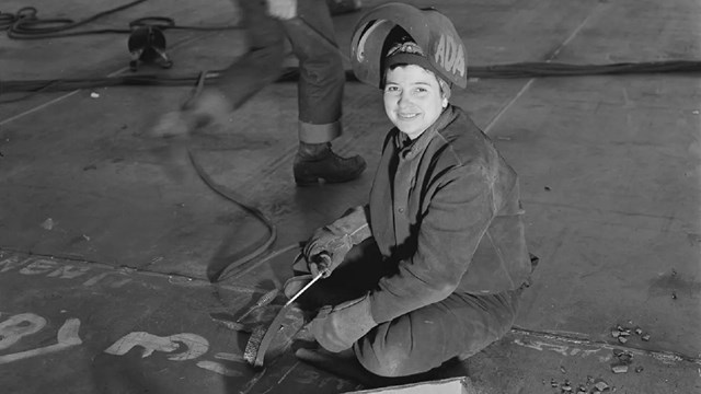 A woman in safety gear smiles at the camera while holding welding equipment