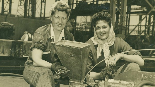 Two women in factory kneeling with machinery; B&W photo