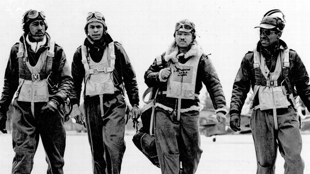 4 african american men in flack jackets and bomber jackets walk along airfield