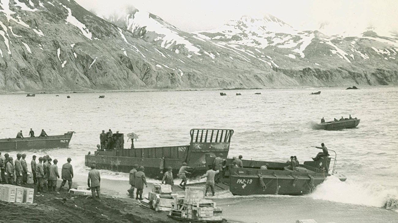 small boats coming ashore, mountains in background; B&W photo