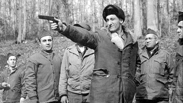 a man stands pointing handgun, with men observing behind him