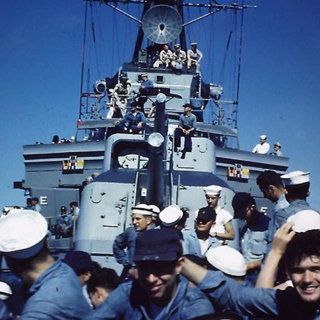 historic photo on deck of battleship facing the 12 inch guns. Sailors are on the deck.