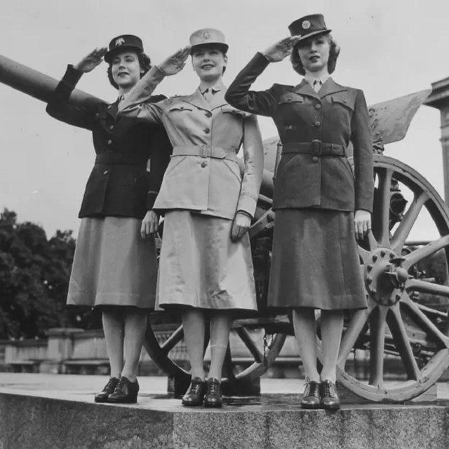World War 2 photo of 3 women in Women's Army Corps uniforms standing in front of a large gun.