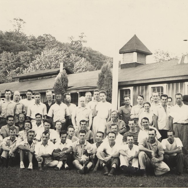 A group of men and a few women pose for a photo outside in front of a low building and trees.
