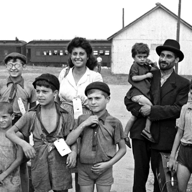 Adults and children with paper tags attached to their clothes stand outside, train in background